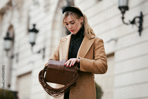 Beautiful young woman opening purse bag and taking something out while standing on street against of building. Stylish female model with fashionable hairstyle