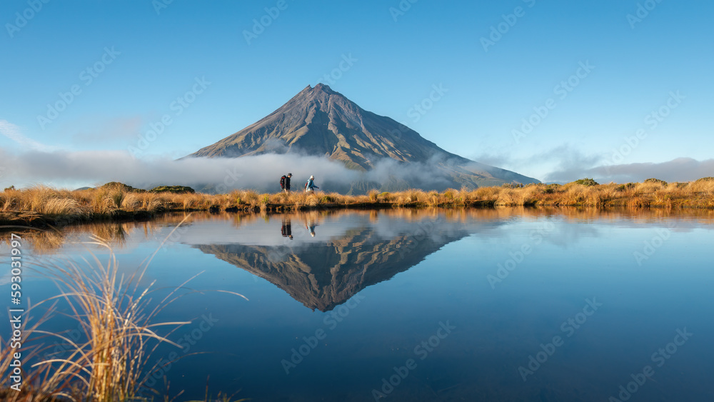 Fototapeta premium Couple hiking Pouakai circuit. Mt Taranaki reflected in the tarn in early morning sunshine. New Zealand.