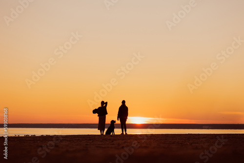 silhouette of people at sunset with dog  two women walking with pet Labrador retriever on the seashore. rest and freedom  view of nature and sunset on sea. poster postcard