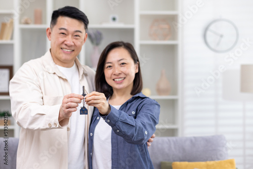 Happy young Asian family  man and woman standing hugging in house  apartment and holding keys in hands  pointing at camera  smiling.