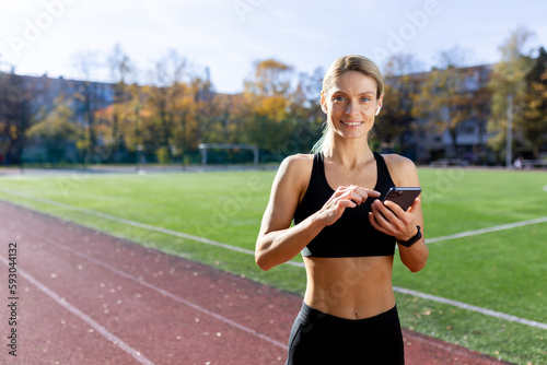 Fototapeta Naklejka Na Ścianę i Meble -  Young female sports coach standing in stadium and using phone, looking at camera smiling.