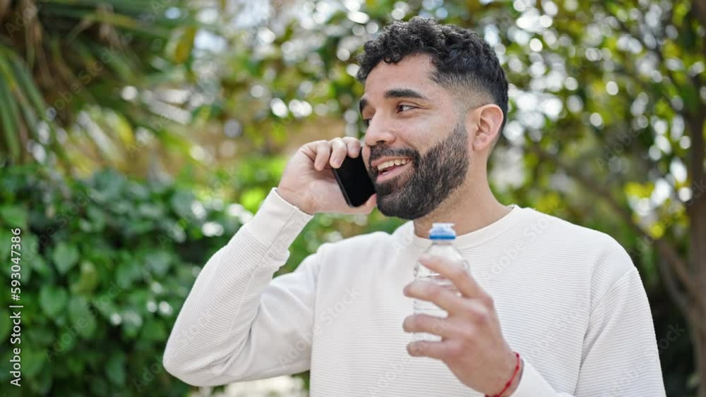 Young hispanic man holding bottle of water talking on smartphone at park Young hispanic man holding bottle of water talking on smartphone at park