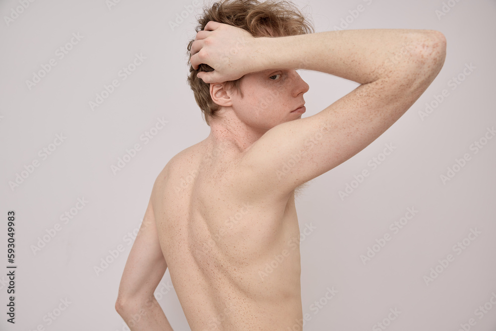 Horizontal back view portrait of a naked young red haired man with freckles on his body showing his body and muscles and lifting his arm up over grey background in studio. Body positive concept. Horizontal back view portrait of a naked young red haired man with freckles on his body showing his body and muscles and lifting his arm up over grey background in studio. Body positive concept.