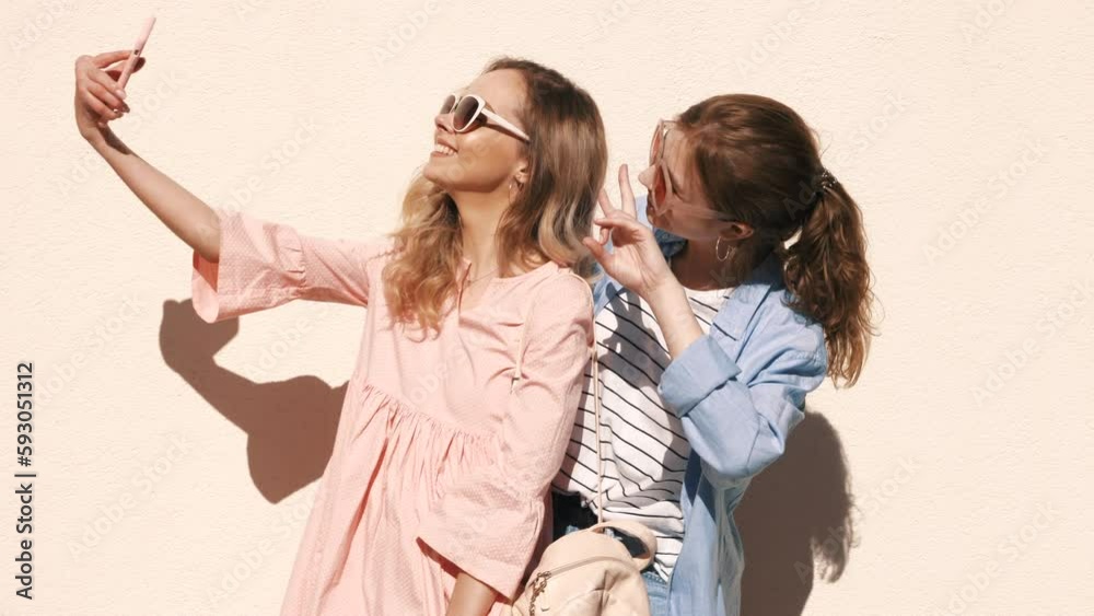 Two young smiling hipster blond women in summer clothes. Girls taking selfie self portrait photos on smartphone. Models posing on street background. Female showing positive emotions Two young smiling hipster blond women in summer clothes. Girls taking selfie self portrait photos on smartphone. Models posing on street background. Female showing positive emotions