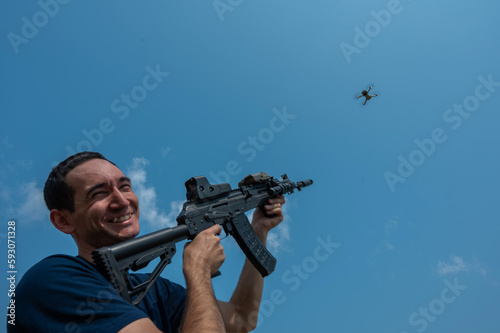 A man aims to shoot a rifle at a flying drone against a blue sky. 
