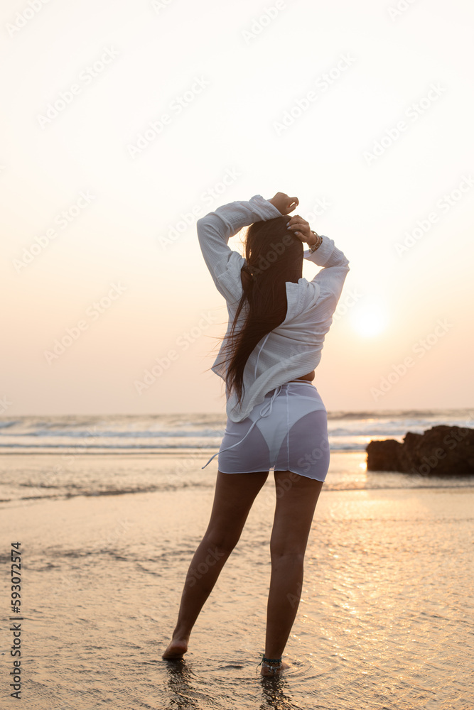 Rearview of sensual brunette long haired woman in wet transparent dress, in bikini swimsuit stand in sea water at resort