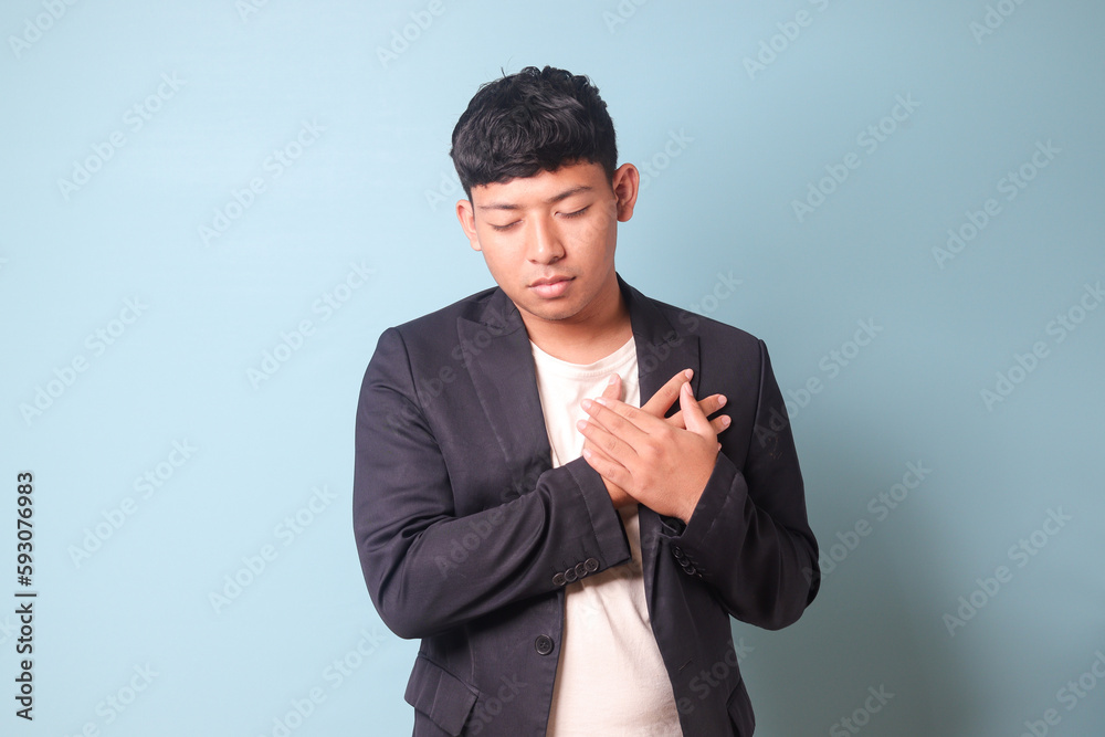 Portrait of young Asian business man in casual suit heart broken holding chest with both hands. Isolated image on blue background Portrait of young Asian business man in casual suit heart broken holding chest with both hands. Isolated image on blue background