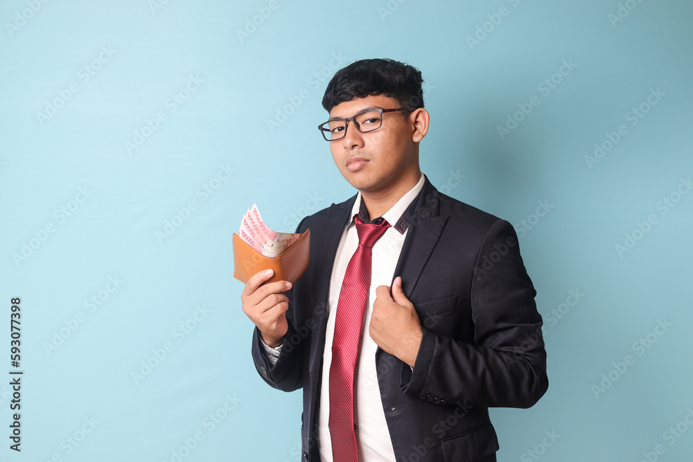 Portrait of young Asian business man in casual suit with confidence expressions fixing collar and holding leather wallet with thousand rupiahs. Isolated image on blue background Portrait of young Asian business man in casual suit with confidence expressions fixing collar and holding leather wallet with thousand rupiahs. Isolated image on blue background