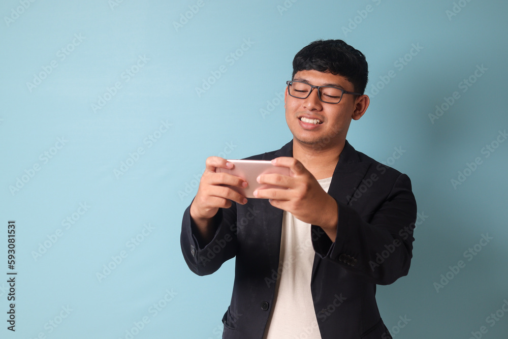 Portrait of young Asian business man in casual suit with big smile looking at phone. Isolated image on blue background Portrait of young Asian business man in casual suit with big smile looking at phone. Isolated image on blue background