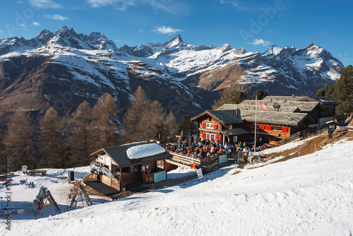 Male and female tourists outside cafe at ski resort with beautiful view of snowcapped mountains and sky in background at Zermatt, Switzerland, winter holiday travel concept