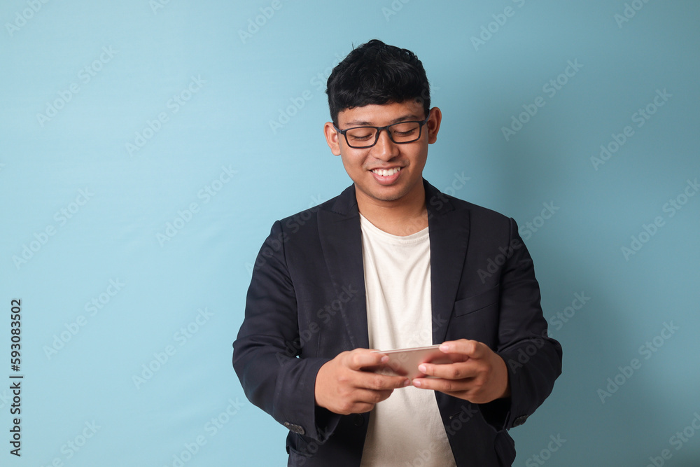 Portrait of young Asian business man in casual suit smiling while holding phone. Isolated image on blue background