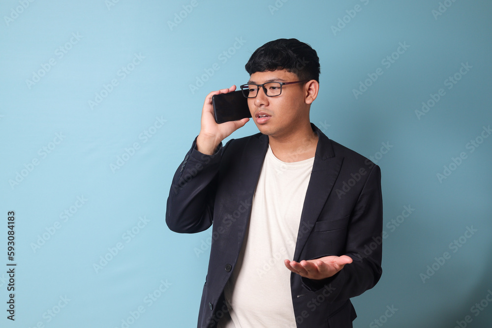 Portrait of young Asian business man in casual suit arguing with someone on the phone. Isolated image on blue background Portrait of young Asian business man in casual suit arguing with someone on the phone. Isolated image on blue background