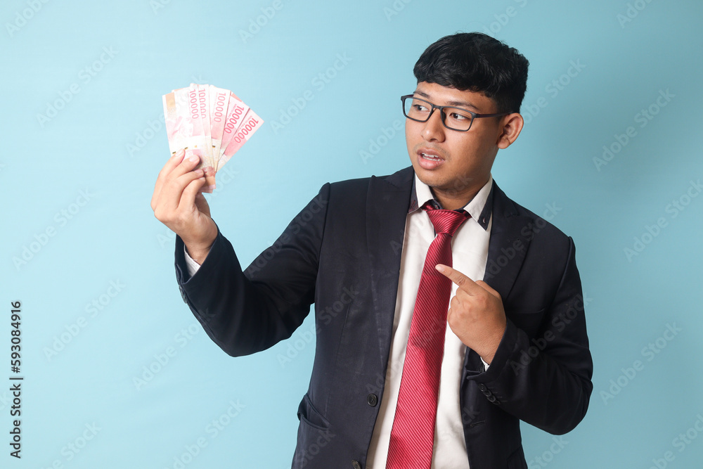 Portrait of young Asian business man in casual suit looking surprised and pointing at thousand rupiahs while holding it. Isolated image on blue background Portrait of young Asian business man in casual suit looking surprised and pointing at thousand rupiahs while holding it. Isolated image on blue background