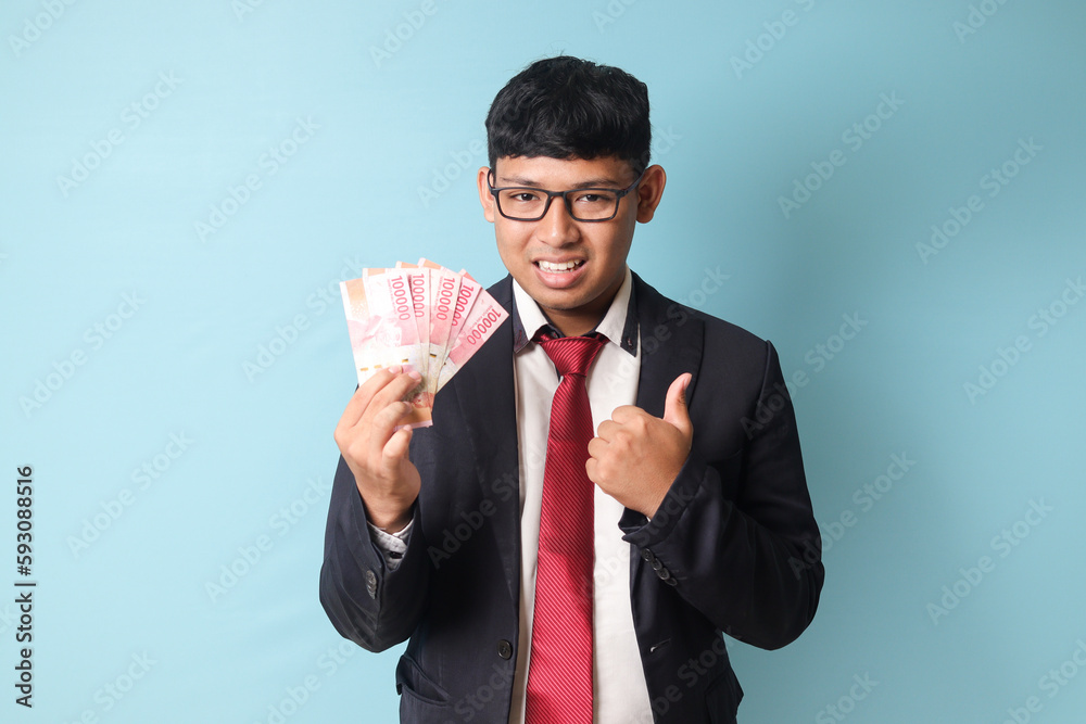 Portrait of happy young Asian business man in casual suit holding thousand rupiahs while making thumbs up. Isolated image on blue background Portrait of happy young Asian business man in casual suit holding thousand rupiahs while making thumbs up. Isolated image on blue background