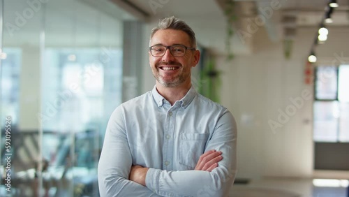 Camera tracks across smiling mature businessman wearing glasses standing in modern open plan office looking into camera - shot in slow motion