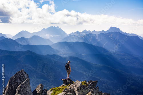 Adventurous athletic male hiker standing on top of a rugged mountain in the Pacific Northwest with jagged mountains in the background.