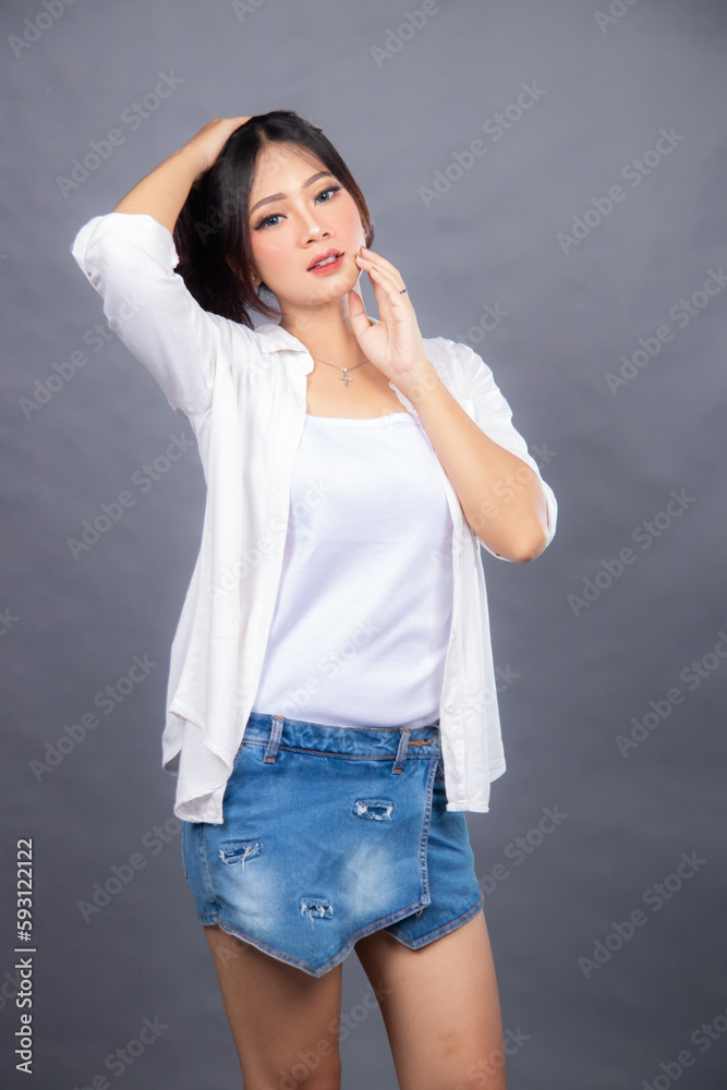 cheerful smiling asian woman posing standing in front of camera holding ladder holding hair and cheeks indoors. fashion promotion women's shirt photo session in gray background studio