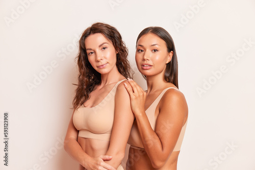 Two pretty seminude girl models in comfortable beige colour bras posing on white background, they stand half-turned one puts hand on other, comfortable underclothing concept, copy space, high quality
