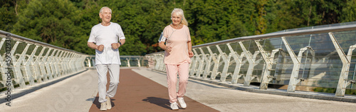 Positive senior gentleman and woman with gadgets run training together along footbridge