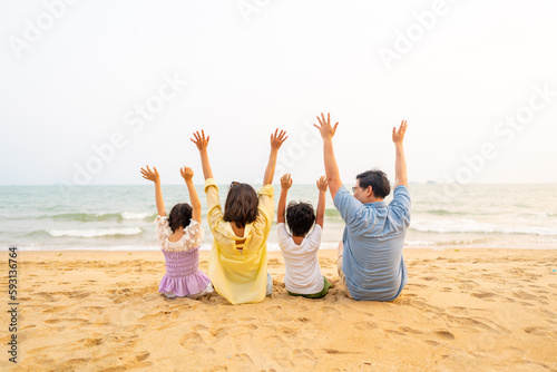Happy Asian family travel ocean on summer holiday vacation. Parents and children kid enjoy and fun outdoor activity lifestyle sitting on tropical island beach with arm raised together at summer sunset