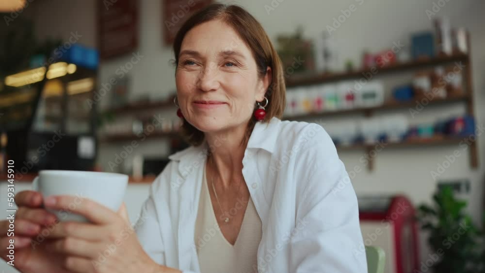Cheerful elderly woman in white shirt drinking tea and looking at camera in cafe Cheerful elderly woman in white shirt drinking tea and looking at camera in cafe