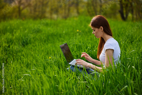 a woman works remotely at a laptop being outdoors in tall grass