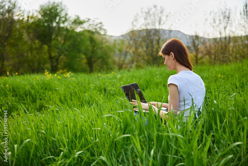 a woman works at a laptop while sitting in high green grass in nature, on a sunny summer day © VICHIZH