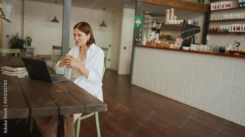 Cheerful mature woman in white shirt talking by video call in headphones on laptop in cafe