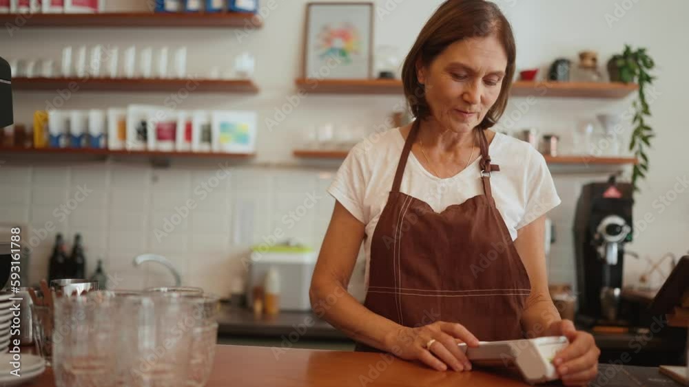 Positive aged woman barista enters amount on terminal and looks into camera behind counter in cafe Positive aged woman barista enters amount on terminal and looks into camera behind counter in cafe