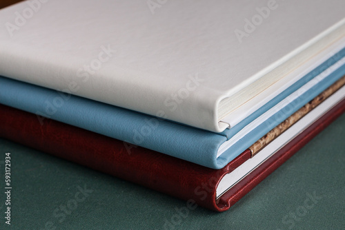Three stylish photobooks with leather covers, white, burgundy and blue, of different thicknesses, lie on a dark green surface in a room.