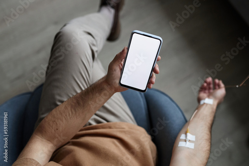 Top view of man using smartphone with white screen mockup during IV drip treatment, copy space