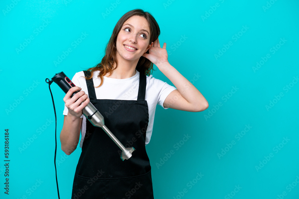 Young caucasian cooker woman using hand blender isolated on blue background listening to something by putting hand on the ear