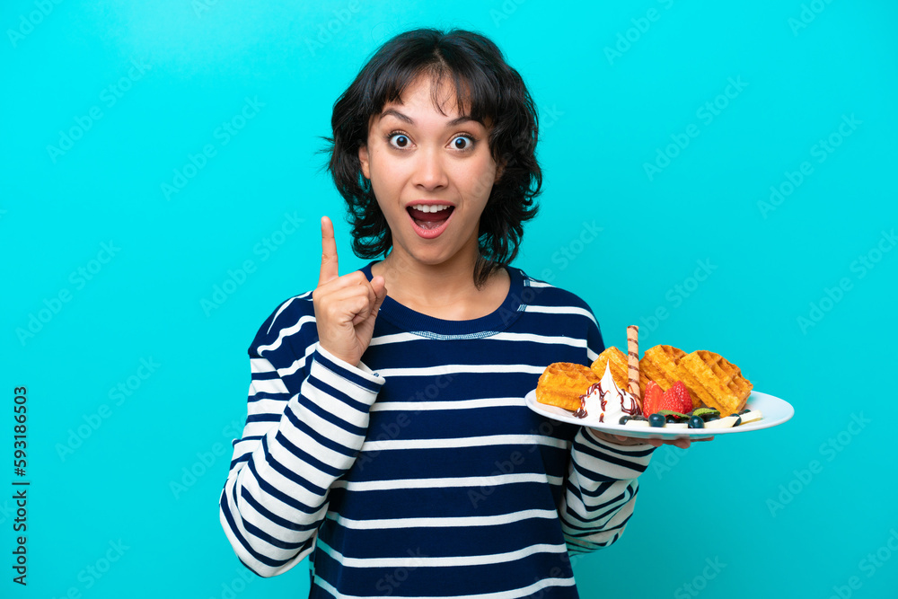 Young Argentinian woman holding waffles isolated on blue background intending to realizes the solution while lifting a finger up Young Argentinian woman holding waffles isolated on blue background intending to realizes the solution while lifting a finger up