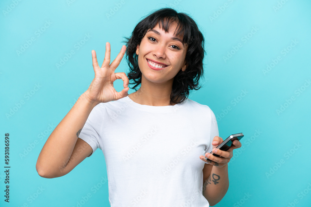 Young Argentinian woman isolated on blue background using mobile phone and doing OK sign Young Argentinian woman isolated on blue background using mobile phone and doing OK sign