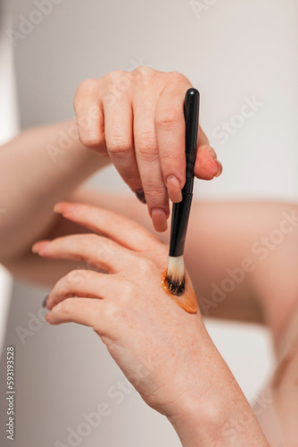 Beautiful young woman holds a makeup brush and applies highlighter cream on her hand  close-up  Beauty and makeup