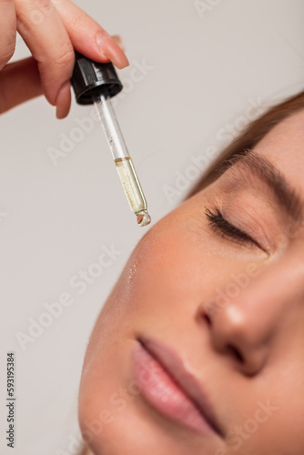 Beautiful young fresh girl drips cosmetic oil from an eyedropper on her facial skin, closeup. Beauty and skin care