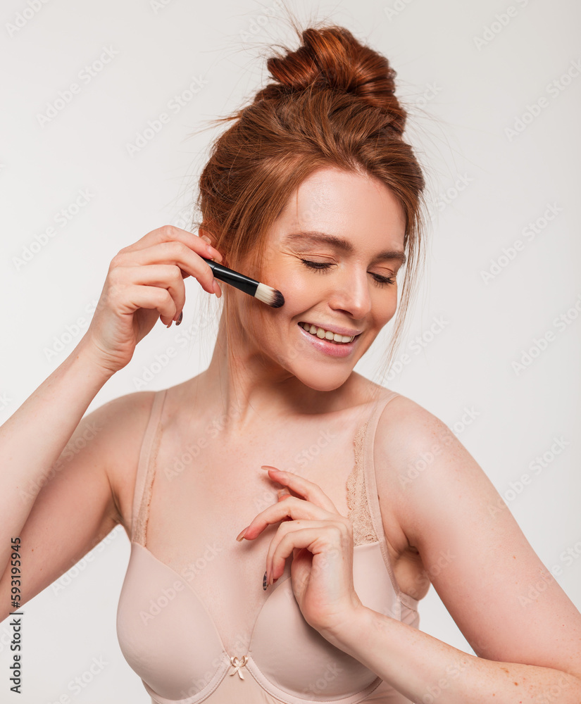 Happy beautiful young fresh redhead woman with a sweet smile doing makeup with a brush in the studio. Beauty and skin care