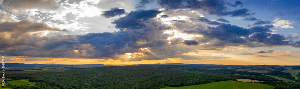 Panoramic Aerial View view of the countryside and cloudy sky with sun rays. Countryside covered with green forest.