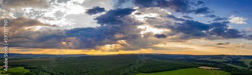 Panoramic Aerial View view of the countryside and cloudy sky with sun rays. Countryside covered with green forest.