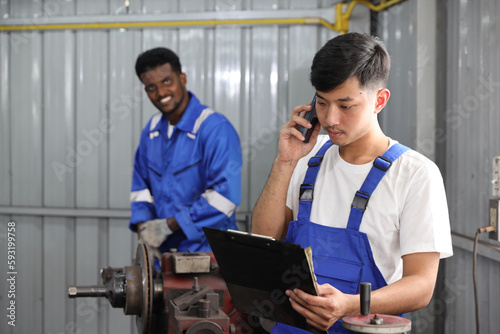 Multiethnic technicain mechanic using metal lathe machine operate polishing car disc brake with smart mobile phone and clipboard at garage. Maintenance automotive and inspecting vehicle part concept