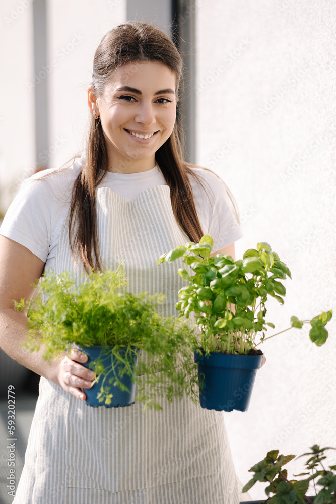 Smiled young female creating garden at balcony. Woman hold pot with dill and basil. Starting for landing