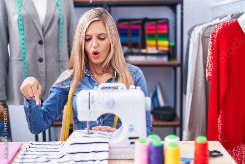Blonde woman dressmaker designer using sew machine pointing down with fingers showing advertisement, surprised face and open mouth