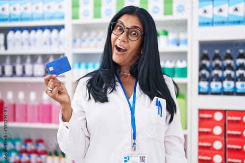 Mature hispanic woman working at pharmacy drugstore holding credit card scared and amazed with open mouth for surprise, disbelief face