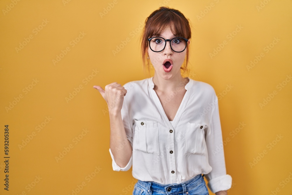 Young beautiful woman wearing casual shirt over yellow background surprised pointing with hand finger to the side, open mouth amazed expression.
