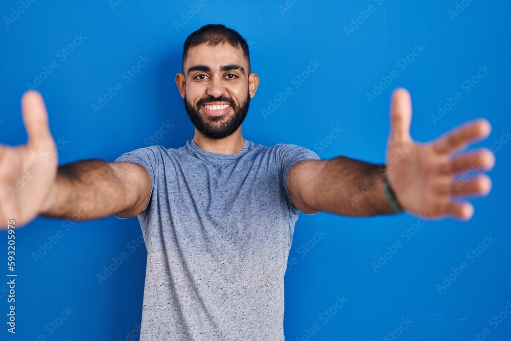 Middle east man with beard standing over blue background looking at the camera smiling with open arms for hug. cheerful expression embracing happiness. Middle east man with beard standing over blue background looking at the camera smiling with open arms for hug. cheerful expression embracing happiness.