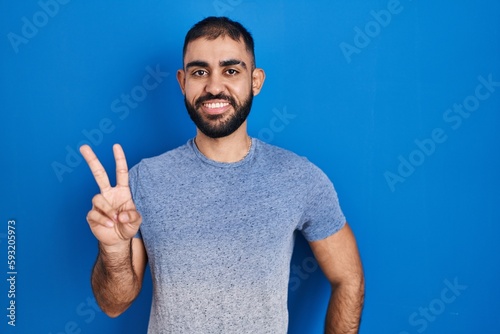 Middle east man with beard standing over blue background showing and pointing up with fingers number two while smiling confident and happy.