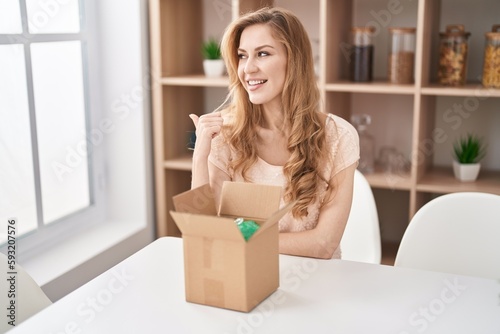 Beautiful blonde woman with cardboard box pointing thumb up to the side smiling happy with open mouth © Krakenimages.com