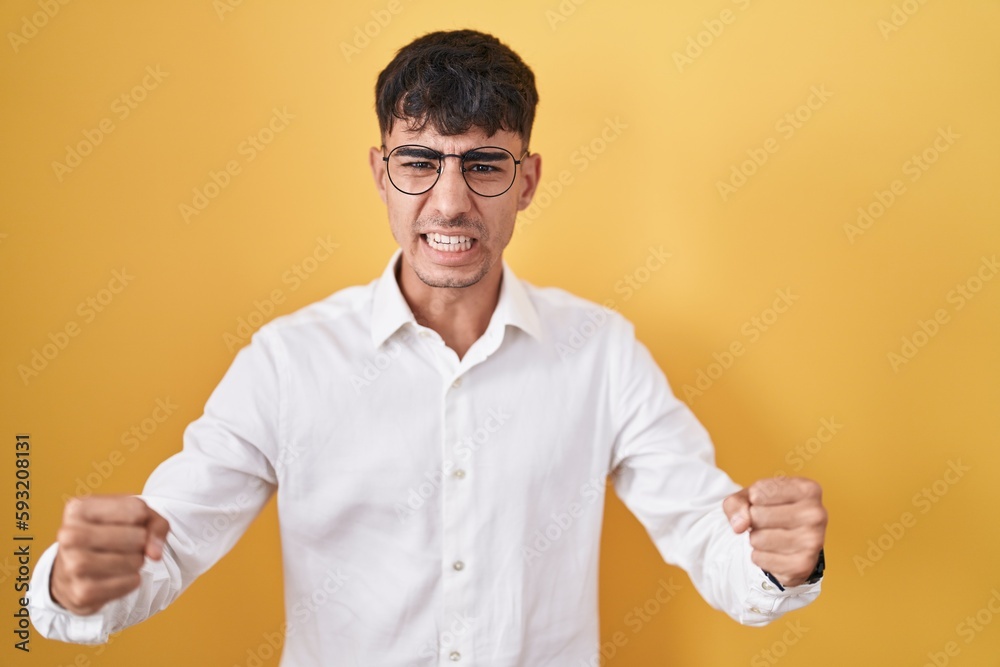 Young hispanic man standing over yellow background angry and mad raising fists frustrated and furious while shouting with anger. rage and aggressive concept.