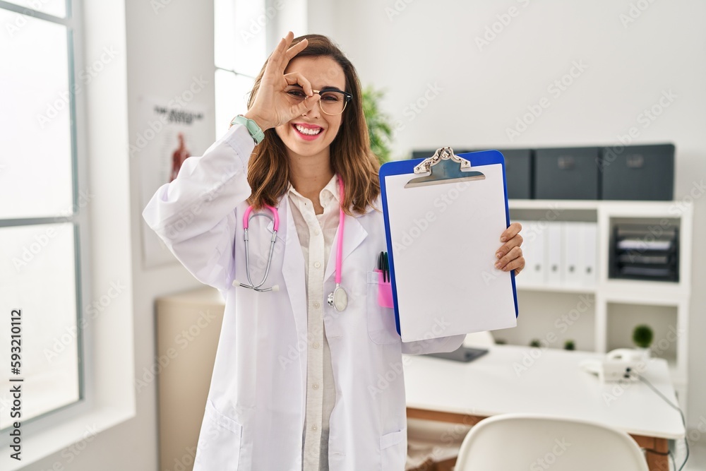 Young doctor woman holding clipboard smiling happy doing ok sign with hand on eye looking through fingers Young doctor woman holding clipboard smiling happy doing ok sign with hand on eye looking through fingers