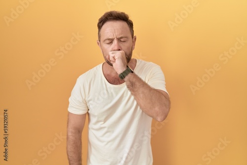 Middle age man with beard standing over yellow background feeling unwell and coughing as symptom for cold or bronchitis. health care concept.